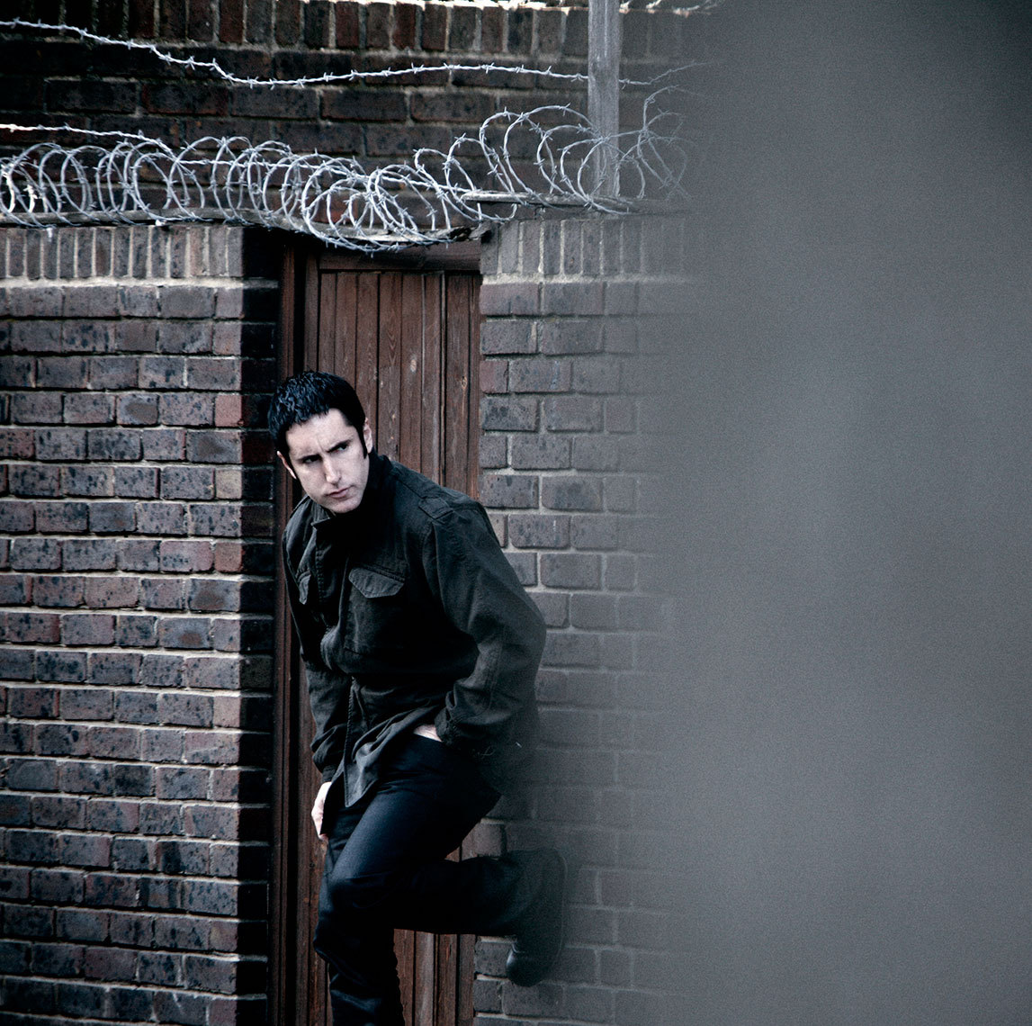 photo of trent reznor standing against a brick wall with barbed wire across the top. He appears to be unaware that he's being photographed